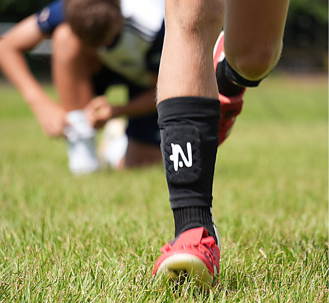 A cinematic close up of a footballer's legs as they run towards the camera wearing the black NTMG sleeved shin pads. In the background the opposing teams player  has stopped chasing and is crouched as they slip their traditional shin pad back in to their sock.