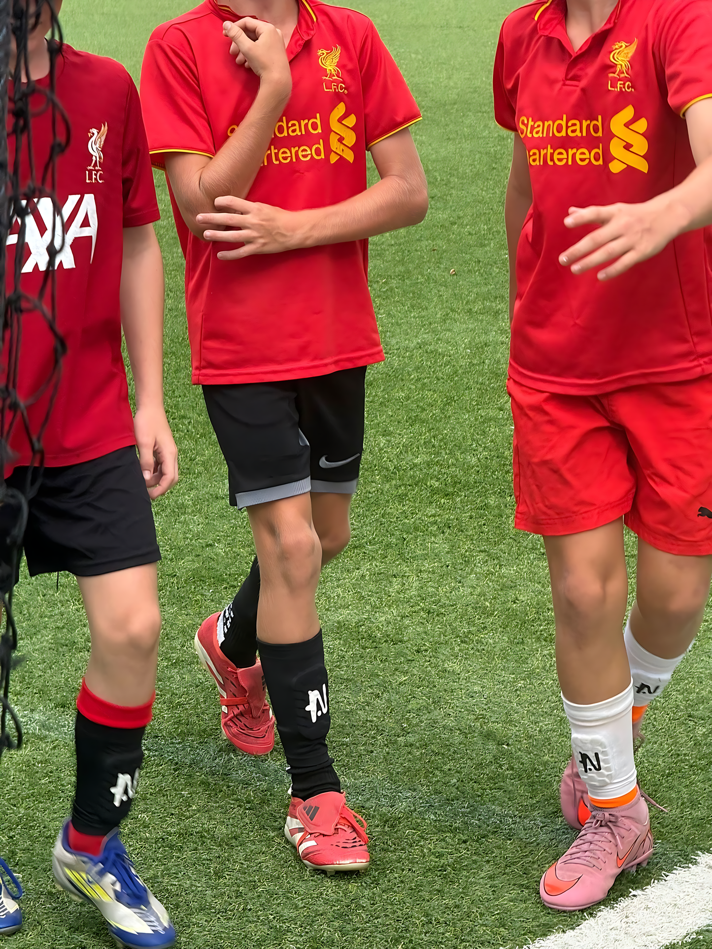 Three kids wearing red sports jerseys with NTMG built in shin guards on a grass field.