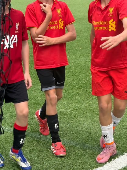 Three kids wearing red sports jerseys with NTMG built in shin guards on a grass field.