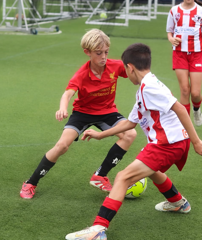 Two children playing soccer on a grass field with one wearing a red jersey and the other in a white and red jersey.