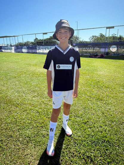 Young boy in sports uniform standing on a grassy field with clear blue sky wearing white NTMG built in shin pads