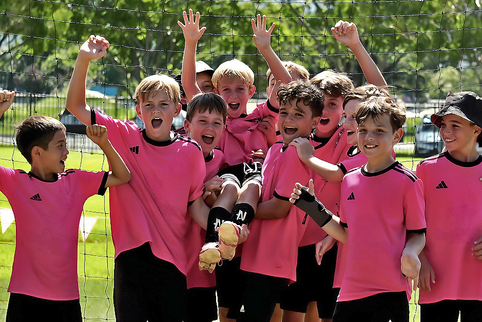 A soccer team of children in pink jerseys celebrate after winning a tournament. The children are holding up a player who is wearing NTMG shin pads.